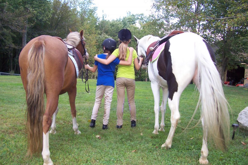 Two young riders at summer camp with arms around each other standing next to a chestnut pony and a black and white paint pony.