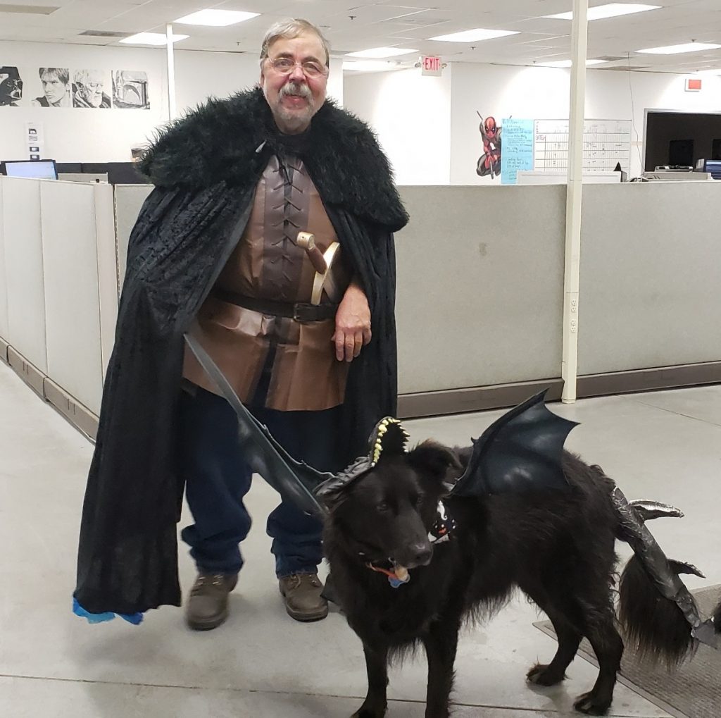 Black dog dressed as a Game of Thrones dragon accompanied by a man dressed in a Jon Snow Halloween costume.
