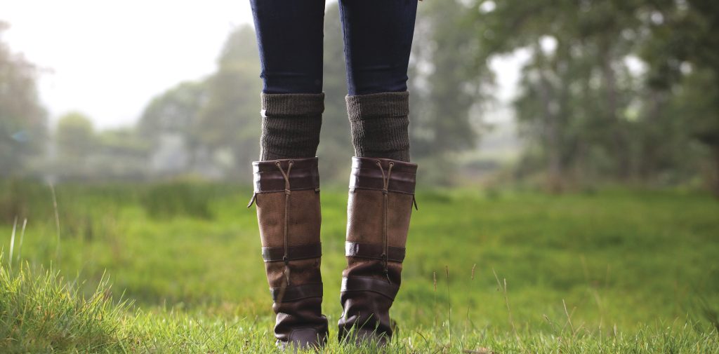 A pair of legs from the thigh down are shown standing in a grassy field. The person is wearing tall country boots over knee-high socks and blue pants.