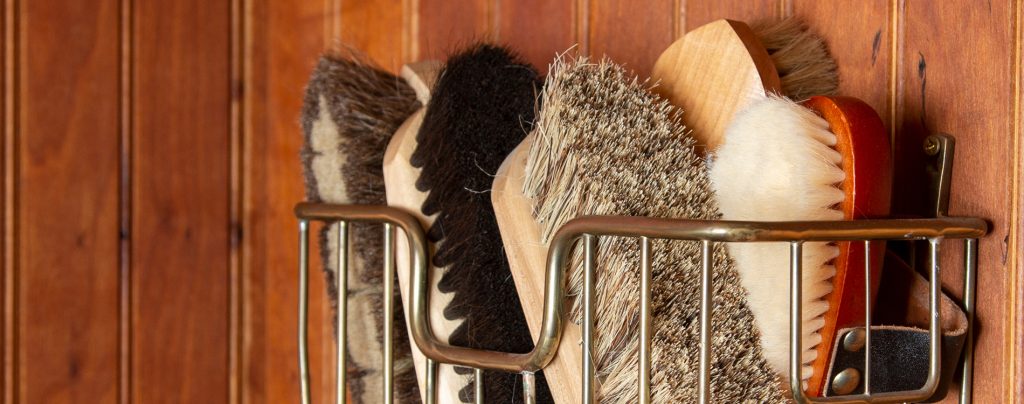 Five brushes are in a metal basket that is hung on a wooden wall. The grooming brushes all have wooden handles with different colored bristles.