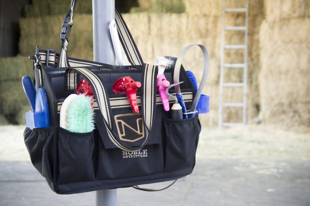 A grooming tote made from black canvas is hanging from a metal pole. There are piles of hay bales in the background and the tote is filled with brushes, curry combs, hoof picks, and spray bottles.