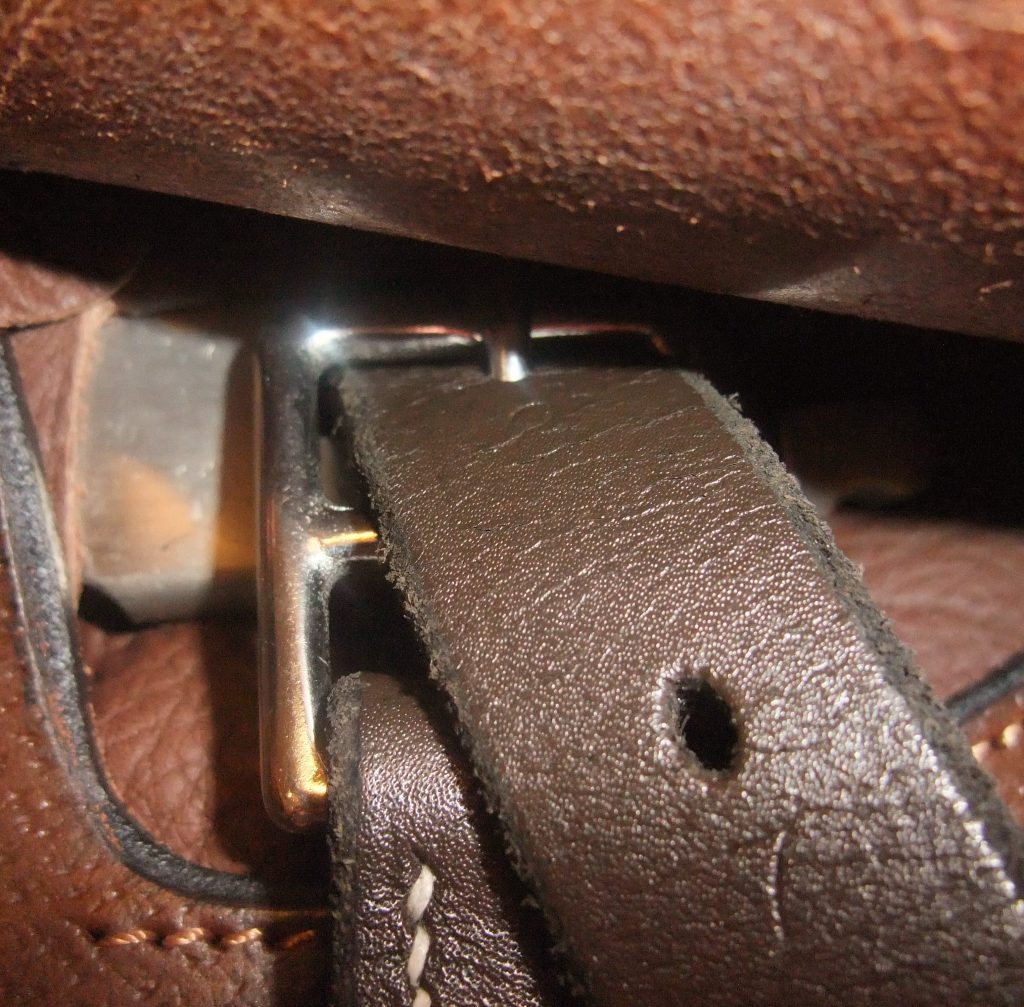 A close-up is shown of a stirrup leather attached to the hook of a saddle underneath the protective flap.