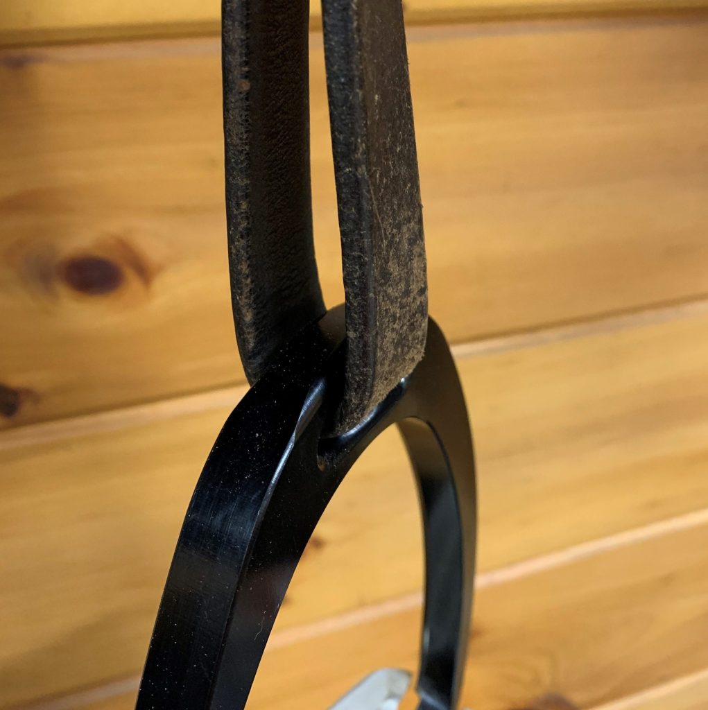 A close-up of a stirrup iron attached to a stirrup leather is shown. The stirrup leather is dark brown, almost black and the stirrup is made of shiny black metal.