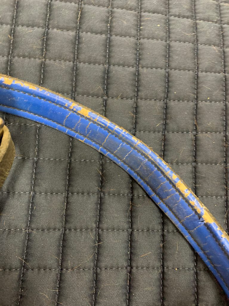 The underside of the crown piece of a blue halter is shown against a dark grey background. The blue leather is cracked and worn.