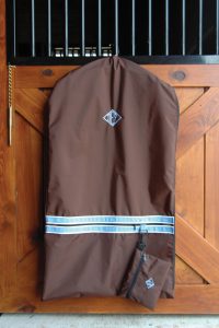 A brown garment bag with light blue accent trim is hanging on the front of a wooden stall door. There is a matching accessory bag attached to the garment bag. Both are embroidered with a diamond with the initials "DEF" inside in light blue.