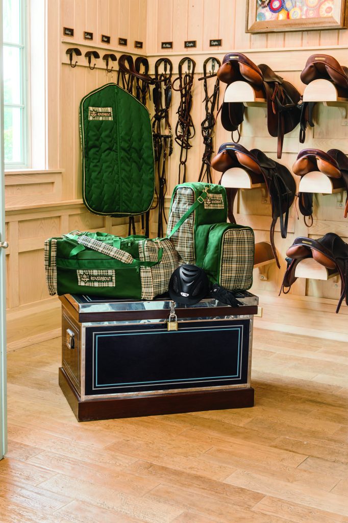 A brightly lit tack room with neat rows of saddles and bridles hung on the wall. There is a navy blue tack trunk and green equipment bags in the middle of the room for storage.