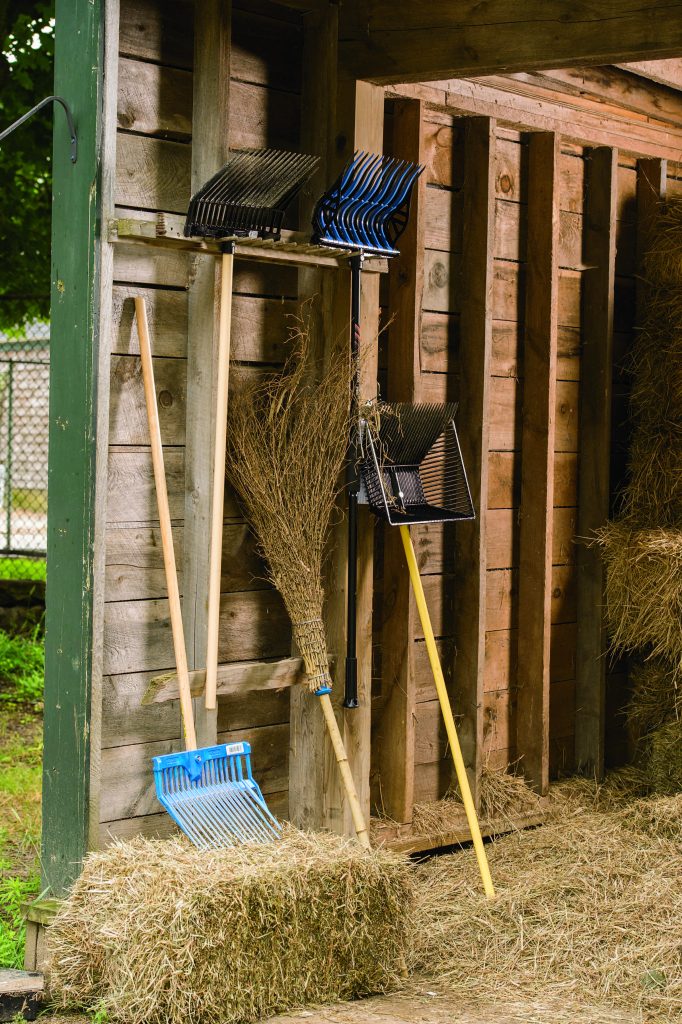 Two pitchforks and one broom are propped up against the wall of a barn. There are also two more pitchforks affixed to the wall. 