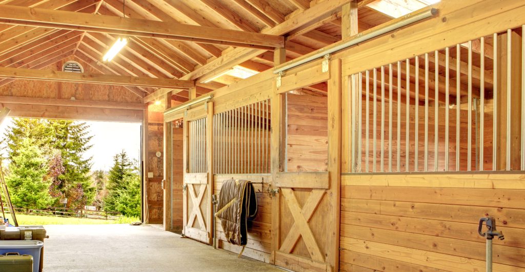 The aisle-way of a clean, brightly lit, horse barn. The barn is made from light colored wood and looks brand new. There is a horse blanket hanging on the wall of one stall and a water spigot in the bottom right hand corner. There are also storage tubs neatly stacked on the left side of the aisle. The barn opens to a wooded area outside.