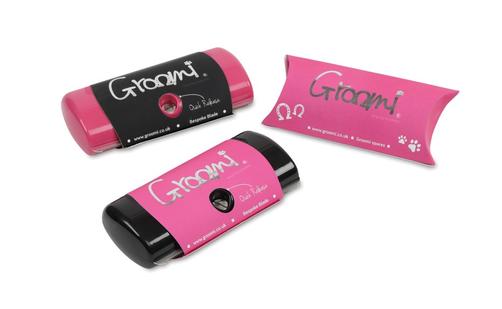 A pink shedding comb with a black label and a black shedding comb with a pink label are pictured and say Groomi. There is also matching pink packaging for the shedding tool next to them.