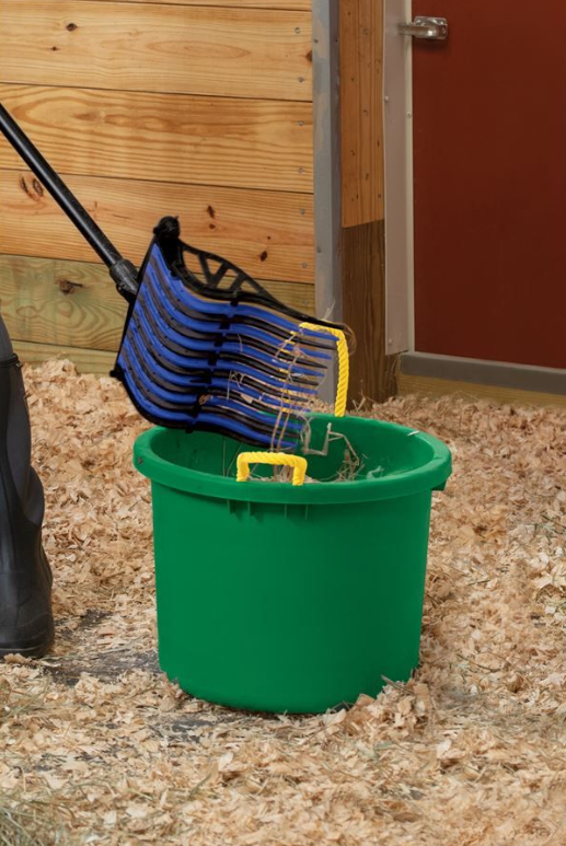 A blue and black pitch fork is being used to dump shavings into a large green plastic bucket with yellow handles.