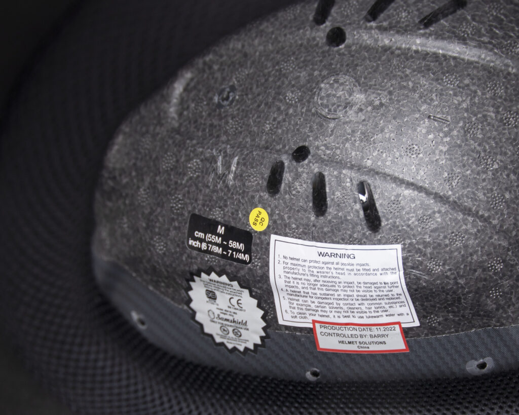 The inside of a Samshield riding helmet shows 5 different stickers regarding the make, model, size and safety information of the helmet. A rectangular white sticker outlined in red with black text notes the production date and series number of this particular helmet.