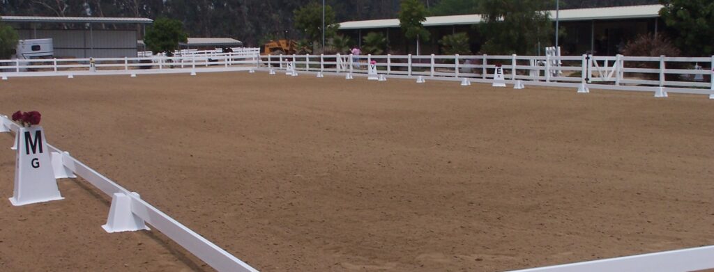 A large outdoor arena with white dressage letter cones and tan arena footing. There is a white fence around the perimeter of the fence.