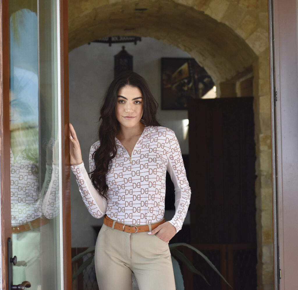 A female model with long brown hair is posed in a tall doorway. She is wearing tan riding pants, a light brown belt, and a white long sleeve quarter-zip shirt with a gold horse bit pattern on it.