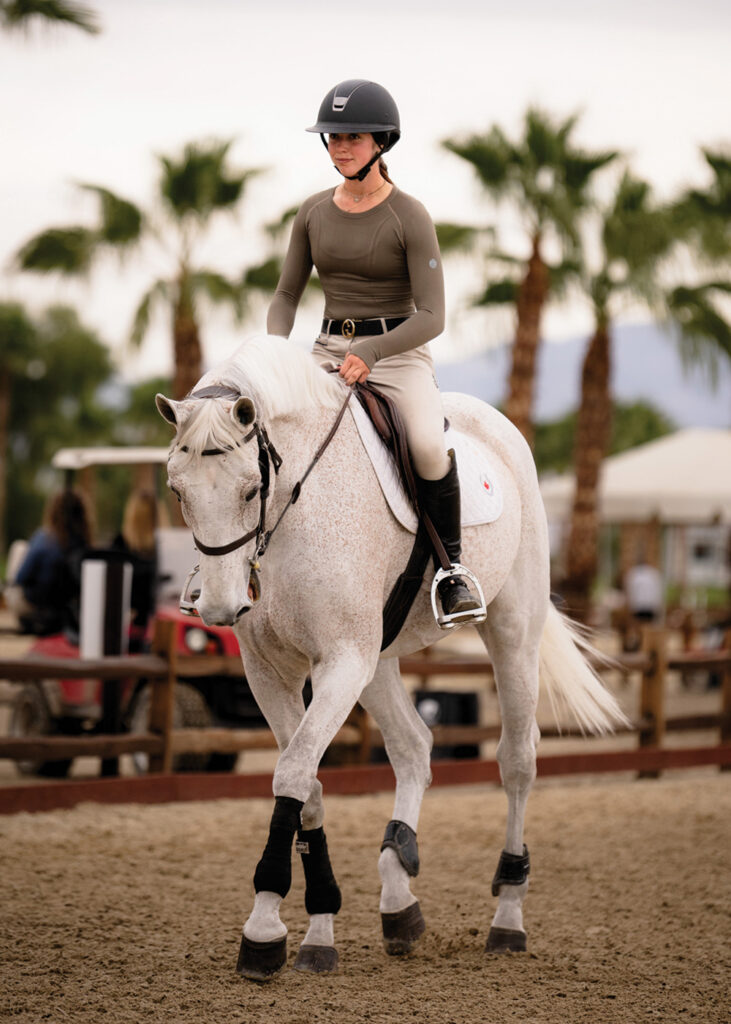A girl wearing tan riding pants and a grey shirt is riding a white horse. They are riding in an outdoor arena with palm trees in the background.