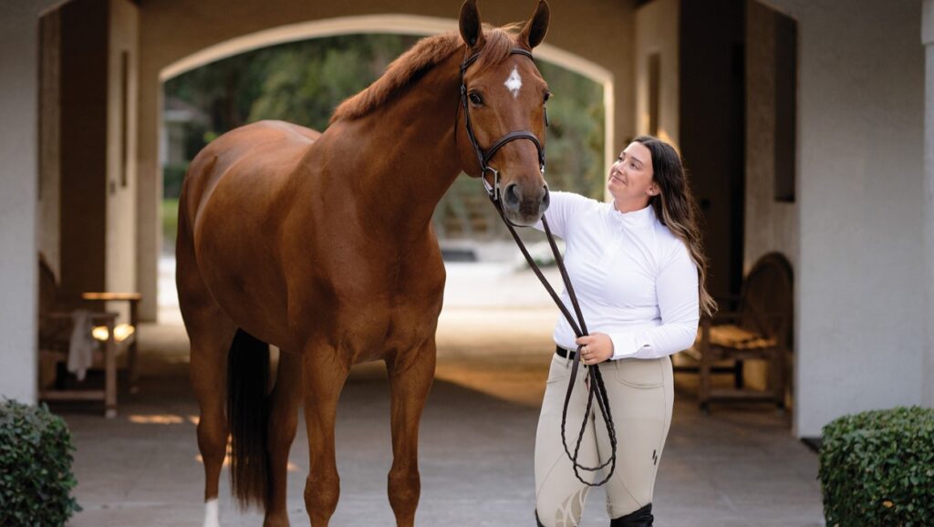 A horse and rider are standing in the hallway of an upscale barn. The rider is a plus size model with long brown hair. She is wearing a long sleeve white show shirt and tan riding pants. She is holding the reins to a large chestnut horse that is only wearing a brown bridle.