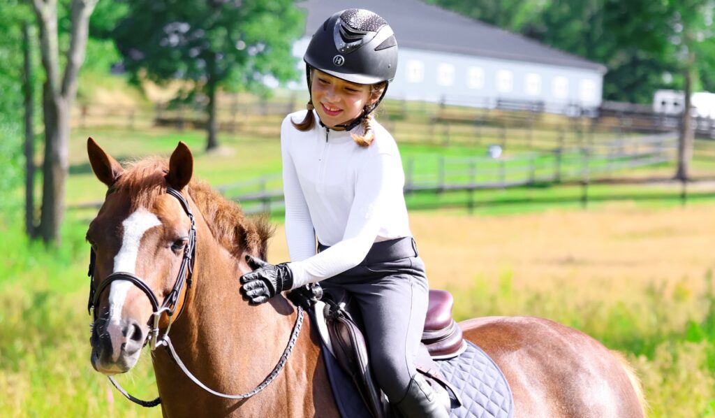 A young girl with brown hair braided into two braids is riding a chestnut pony. The girl is wearing a black helmet, white riding shirt and grey riding tights.