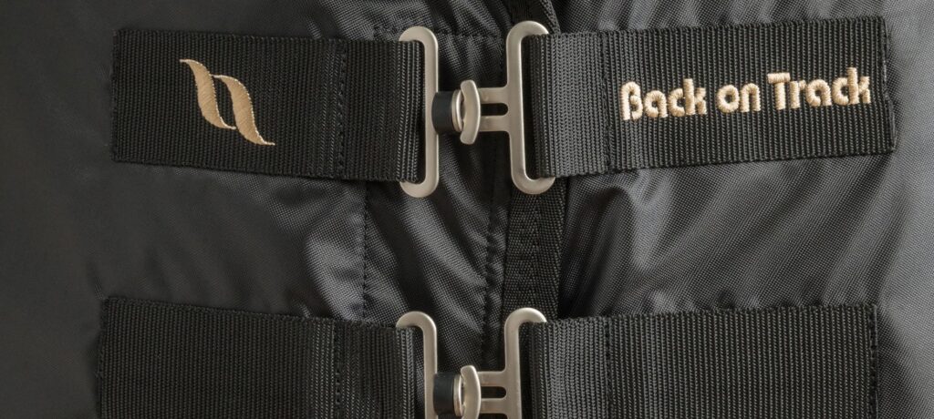 A close up shot of the front chest closures of the Back on Track Royal Mesh Deluxe Sheet in all black. There are two T-Buckles to secure the sheet closed with the top buckle branded with the Back on Track logo in gold thread.