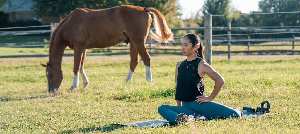 A woman with a long dark brown ponytail, wearing a black tank top and teal yoga leggings is performing a yoga stretch on a yoga mat in a grassy field with a horse in the background.