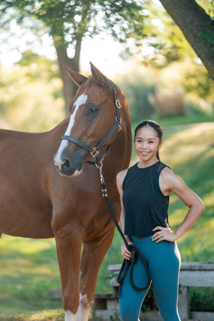 A woman with a long dark brown ponytail, wearing a black tank top and teal yoga leggings is facing the camera holding the lead line to a chestnut colored horse wearing a brown leather halter.