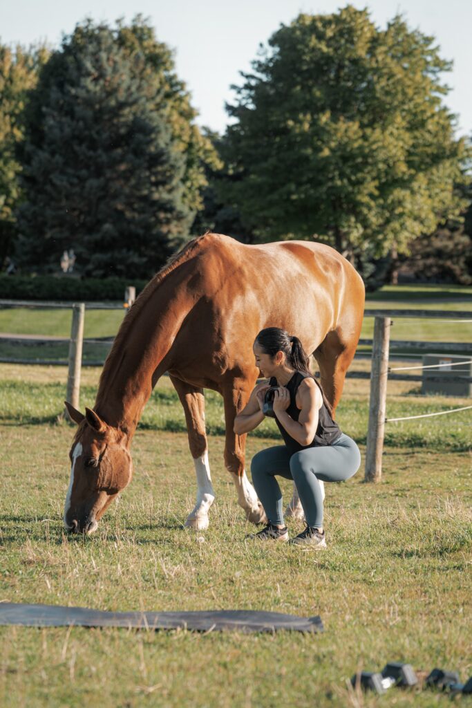 A woman with a long dark brown ponytail, wearing a black tank top and gray yoga leggings is performing a squat in a grassy field next to a horse.