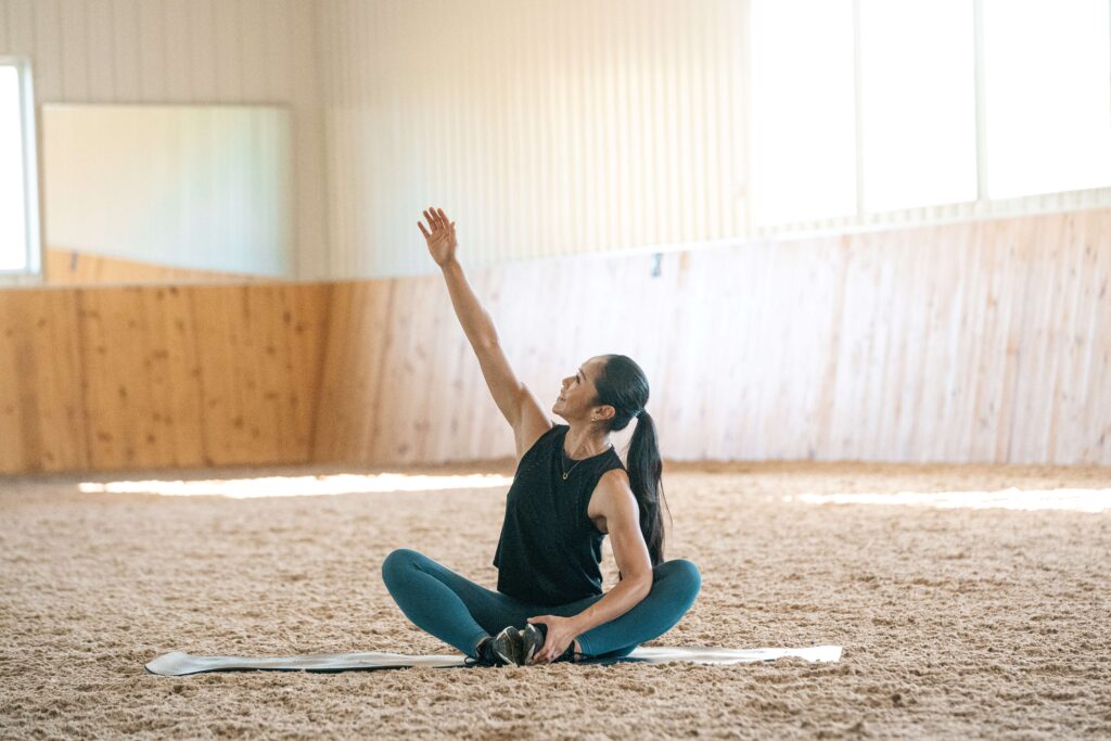 A woman with a long dark brown ponytail, wearing a black tank top and teal yoga leggings is performing a yoga stretch on a yoga mat in a well-lit indoor riding arena.