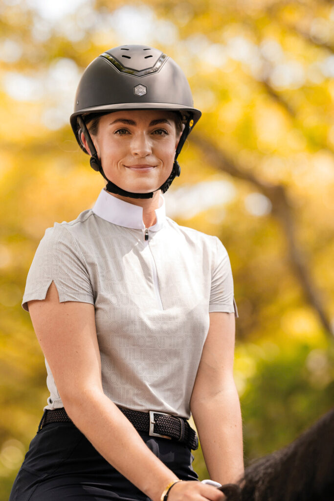 A young woman is seen sitting on a horse that has been cropped out. She is wearing a dark grey riding helmet and a light grey short-sleeve shirt with a white collar. She also has a black belt and black breeches on. She is against a background of trees with yellow fall leaves.
