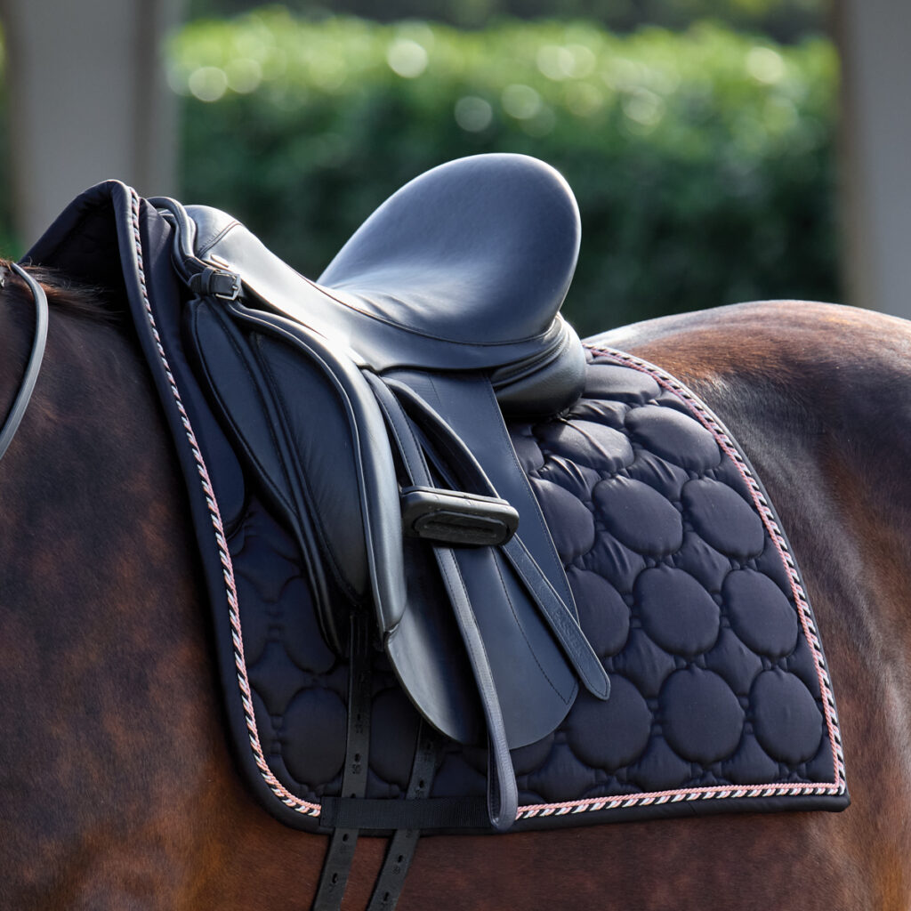 A close up of a dressage saddle with the gift of a Rider's International Luxury saddle pad that is black on a bay horse