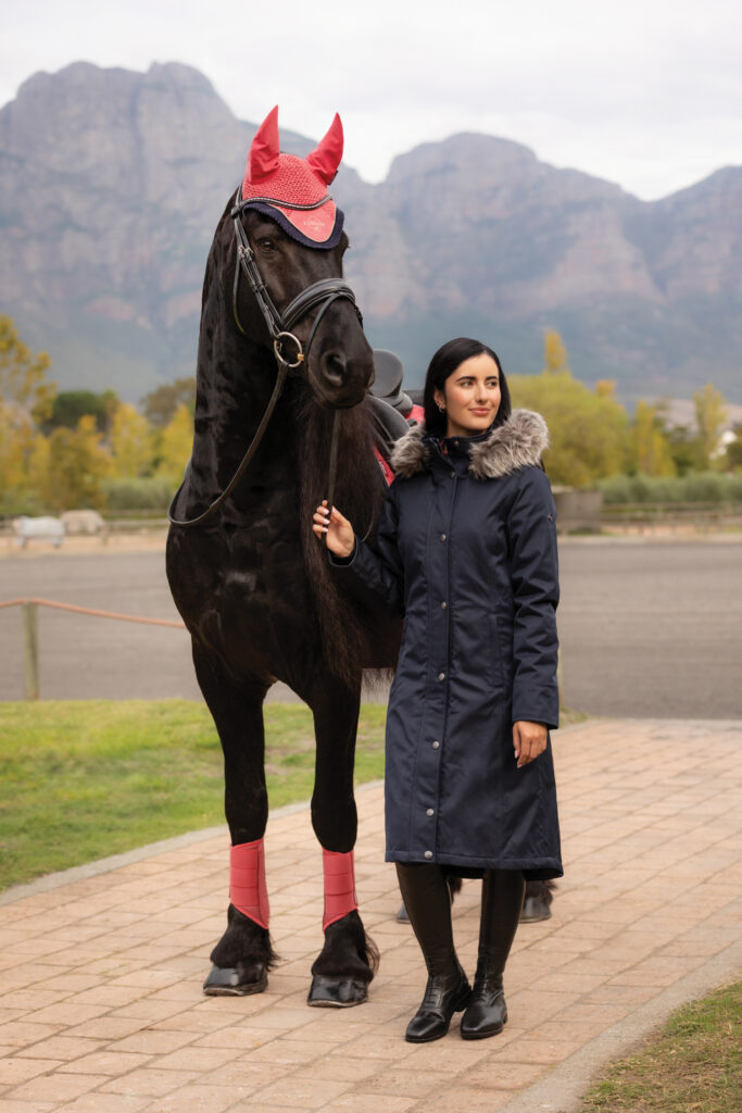 Rider wearing the gift of a Lemiuex long navy coat while holding a black horse with mountains in the background