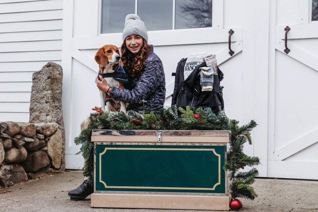 Young rider holding a dog while sitting on a holiday garland on a tack box that is green/tan. A white barn in the background - gift